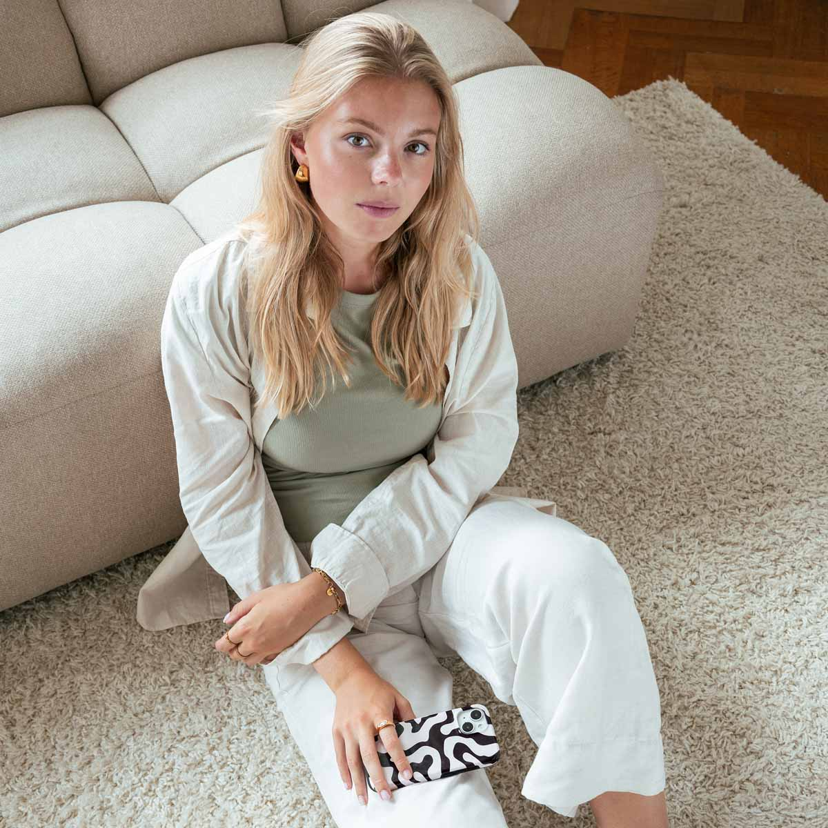 Una donna bionda seduta su un tappeto bianco contro un divano beige, che tiene un telefono fantasia.