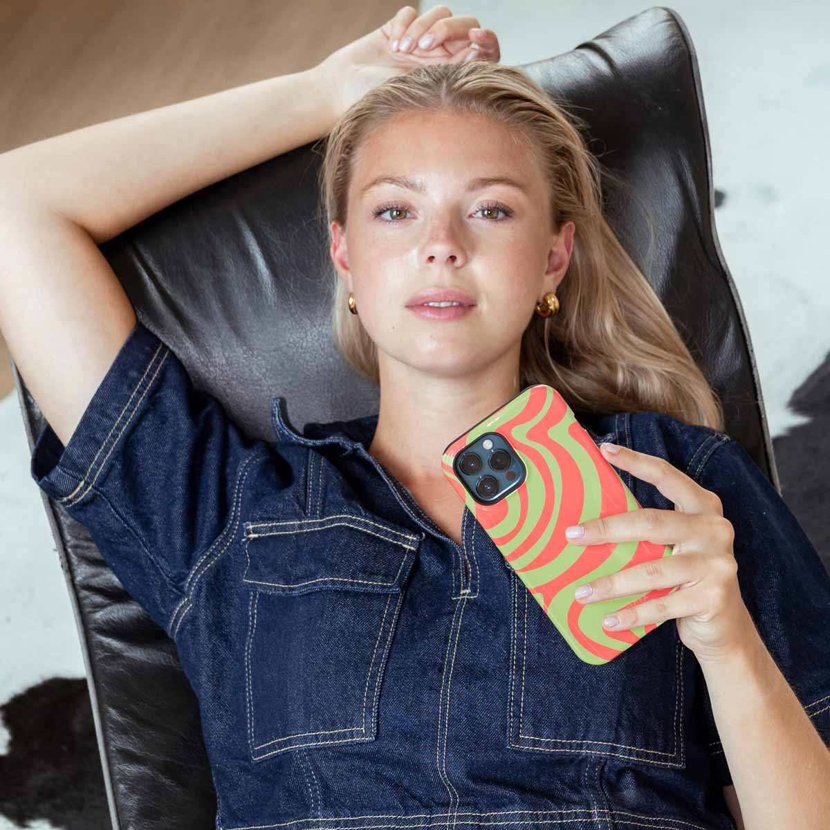 Una mujer rubia con camisa vaquera descansa en una silla oscura sosteniendo un teléfono con funda ondulada roja y verde.