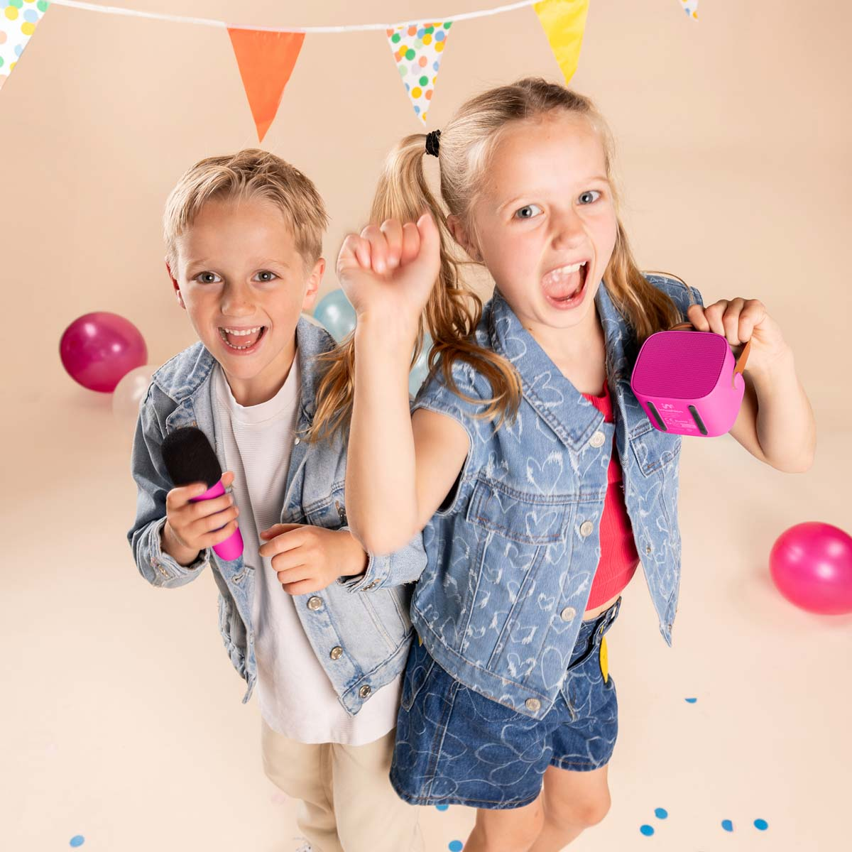 Dos niños cantan y bailan alegremente con un micrófono y un altavoz ante un fondo de fiesta.