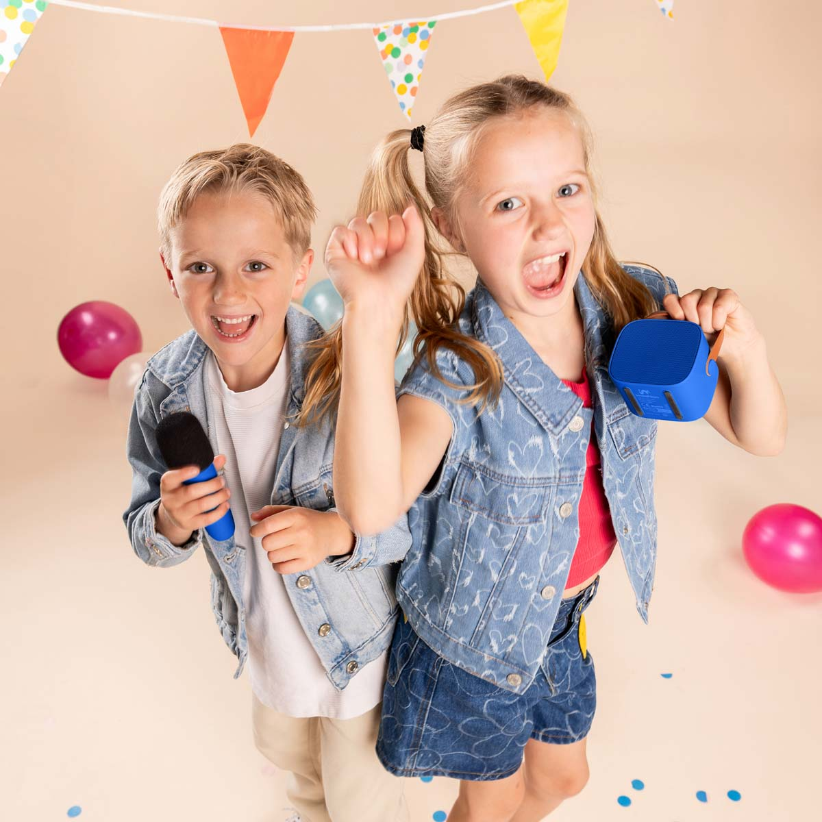Una niña rubia y un niño con micrófono y altavoz portátil, rodeados de globos coloridos y banderines festivos.