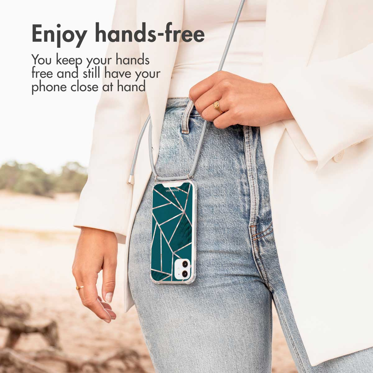 Une personne porte une coque de téléphone bandoulière à motif géométrique vert et or, un jean et un blazer blanc.
