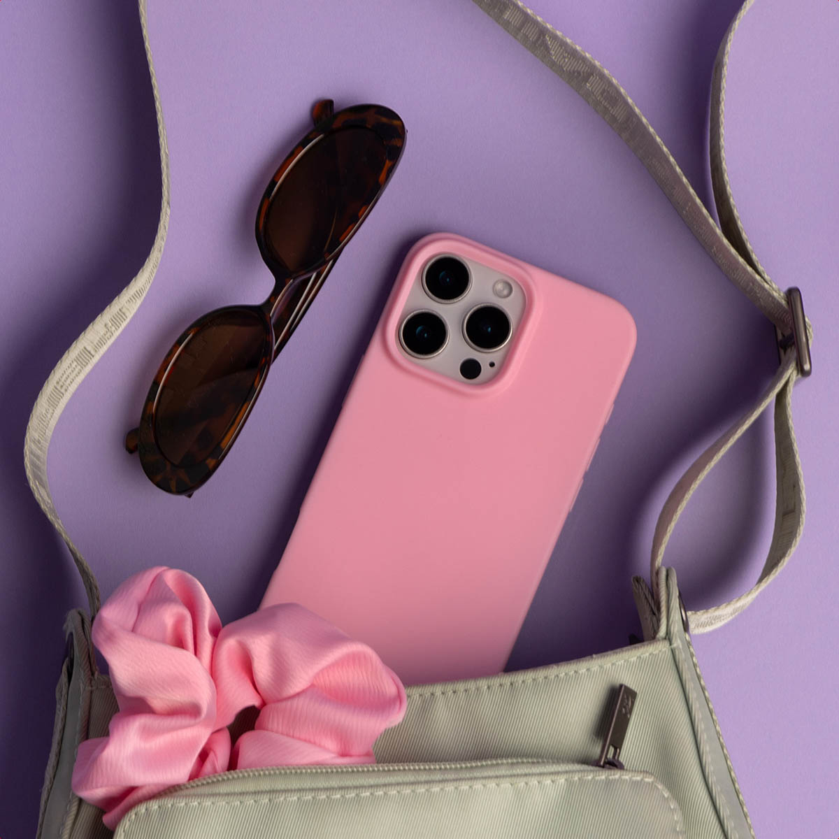Un teléfono rosa, gafas de sol y coletero rosa en un bolso gris sobre morado.