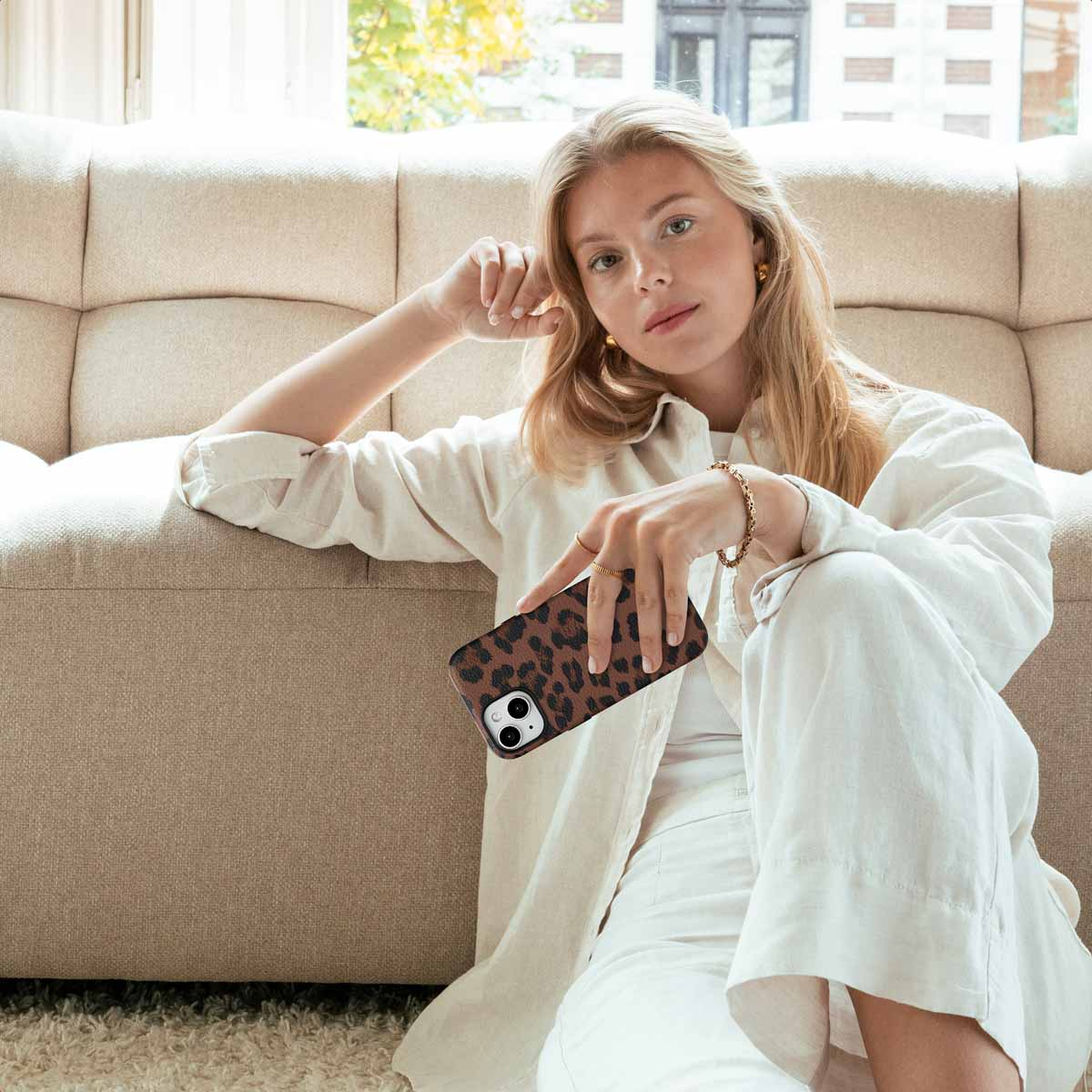 Una donna bionda con camicia bianca siede a terra davanti a un divano, tenendo uno smartphone con stampa leopardata.
