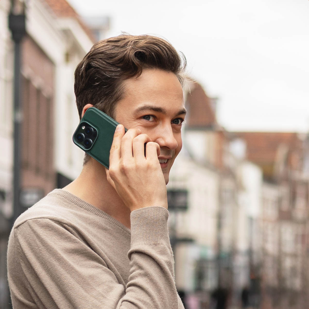 Un giovane con maglione marrone telefona con uno smartphone verde in una strada sfocata del centro storico.