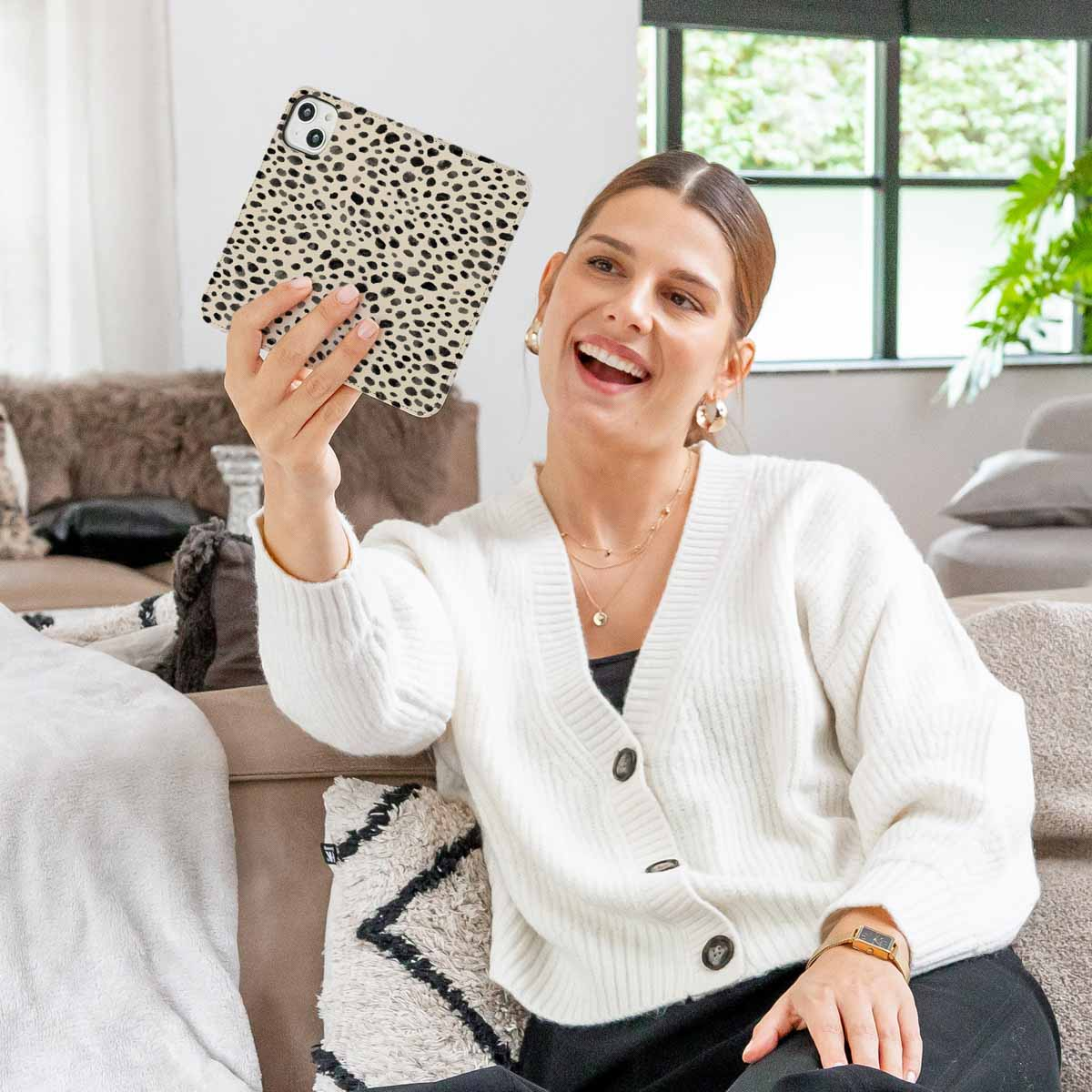 Una mujer sonriente con cárdigan blanco sostiene un teléfono con funda de guepardo en su sala.