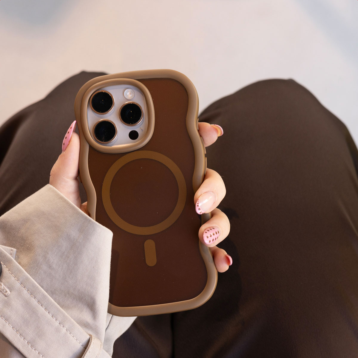 Mano sostiene un smartphone marrón con funda MagSafe, triple cámara y bordes ondulados.