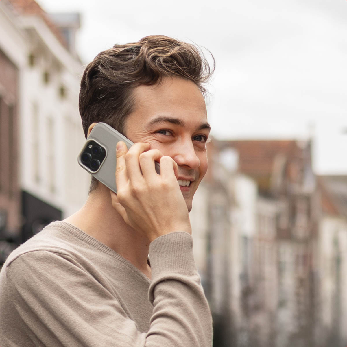 Un joven sonriente habla por teléfono con un smartphone gris frente a un fondo de calle borroso.