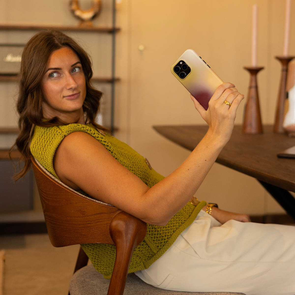 Una donna con un top verde siede a un tavolo di legno e scatta un selfie con lo smartphone.