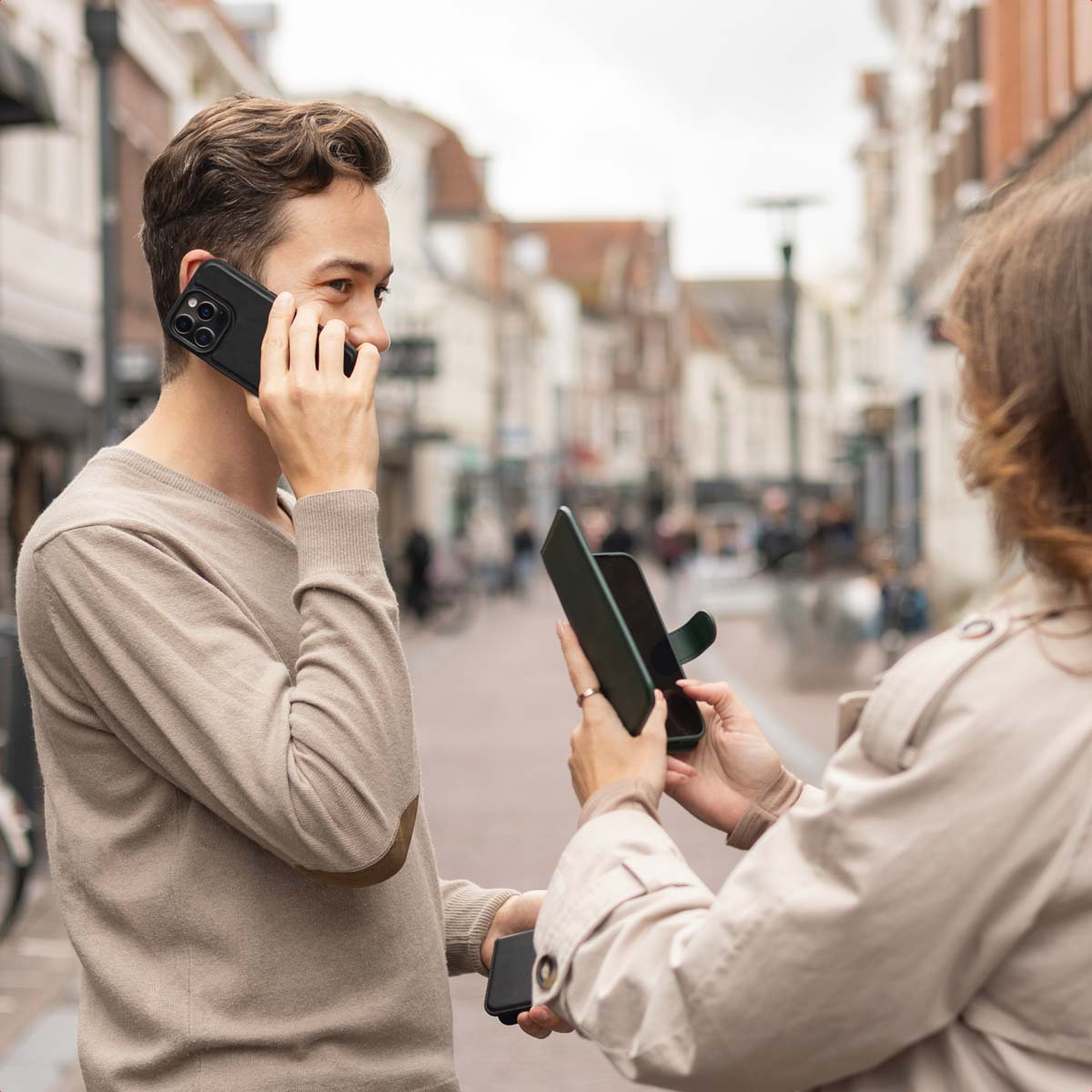 Un uomo con un maglione beige parla al cellulare all'aperto mentre una donna tiene una custodia verde.