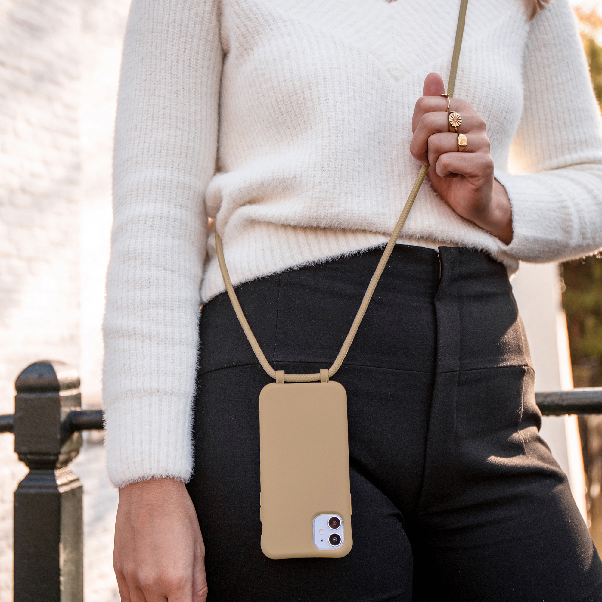 Une femme porte une coque de téléphone beige avec cordon autour du cou, un pull blanc et un pantalon noir.