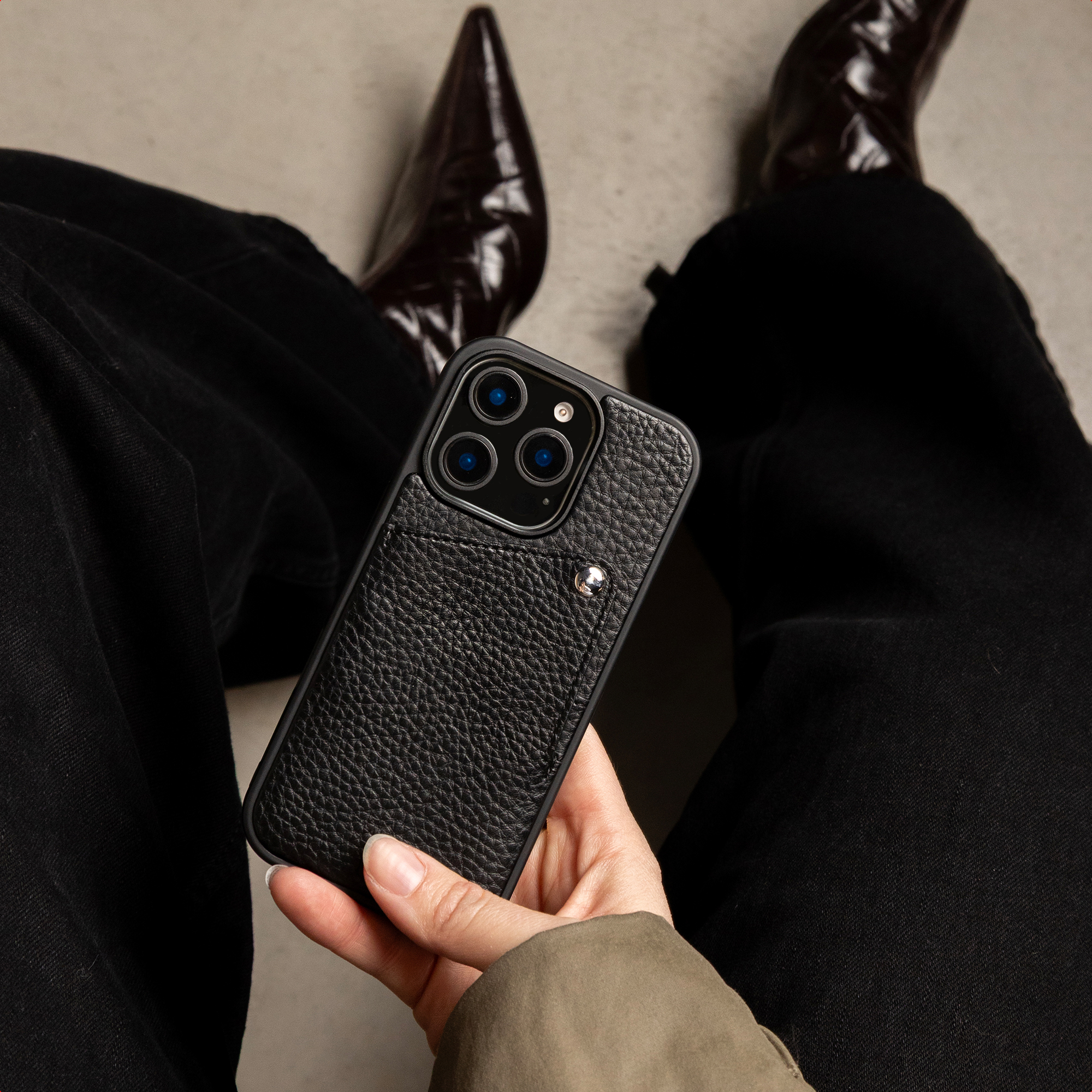 Une main tient une coque de smartphone en cuir noir texturé sur un fond de pantalon noir et de bottes pointues.