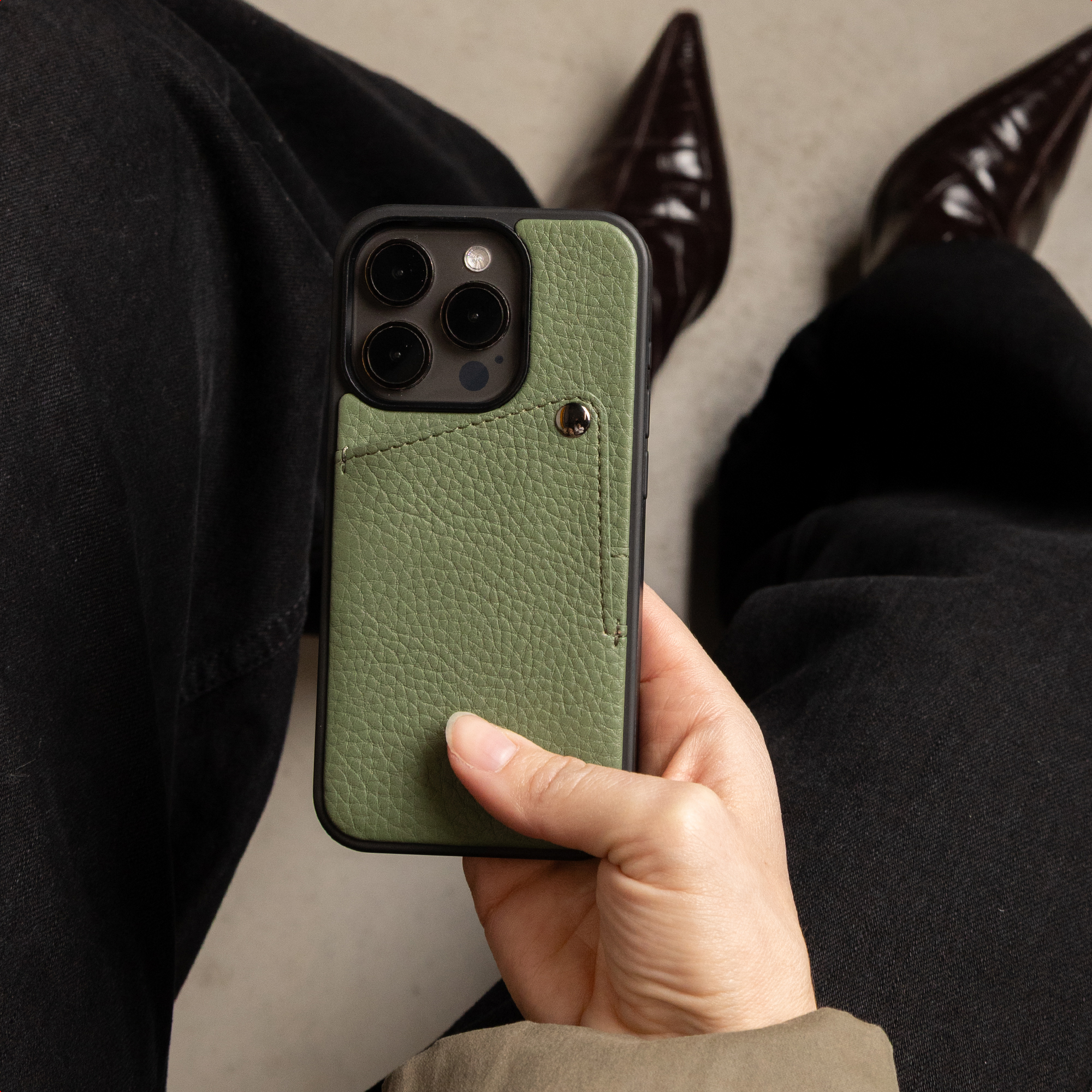Une main tient un smartphone avec une coque en cuir vert texturé sur fond de pantalon noir et chaussures sombres.