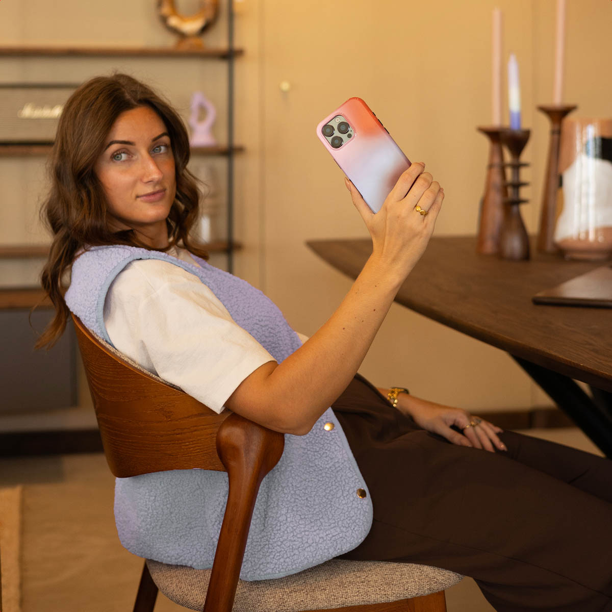 Una donna siede su una sedia sollevando uno smartphone mentre sorride.