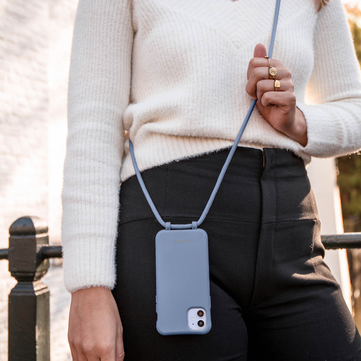Une femme en pull blanc porte un smartphone bleu avec une lanière sur un pantalon noir.