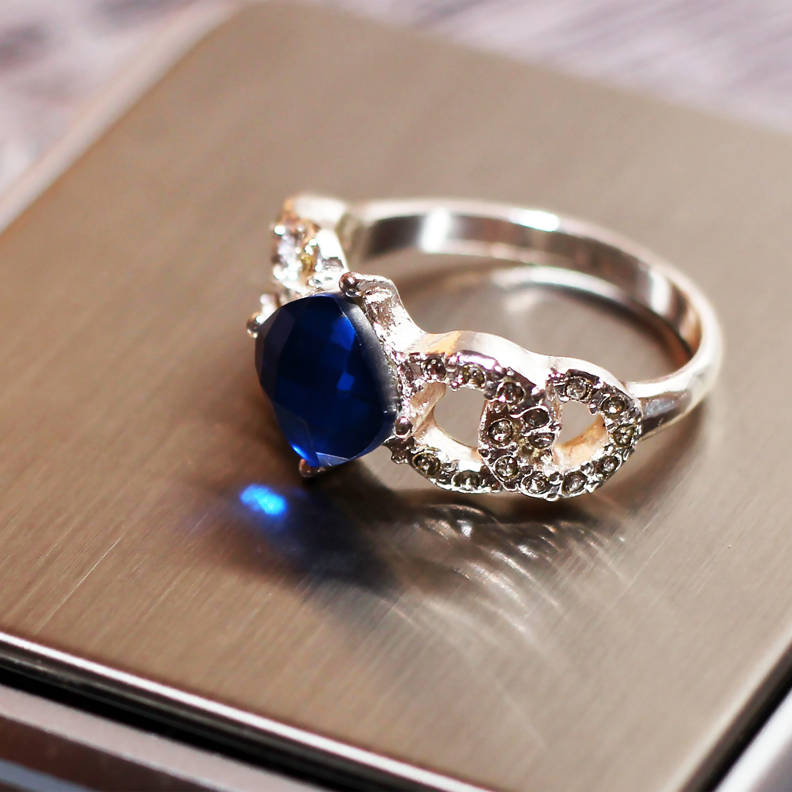 Ein silberner Ring mit einem großen blauen Edelstein und kleinen Strasssteinen auf einer Metallfläche.