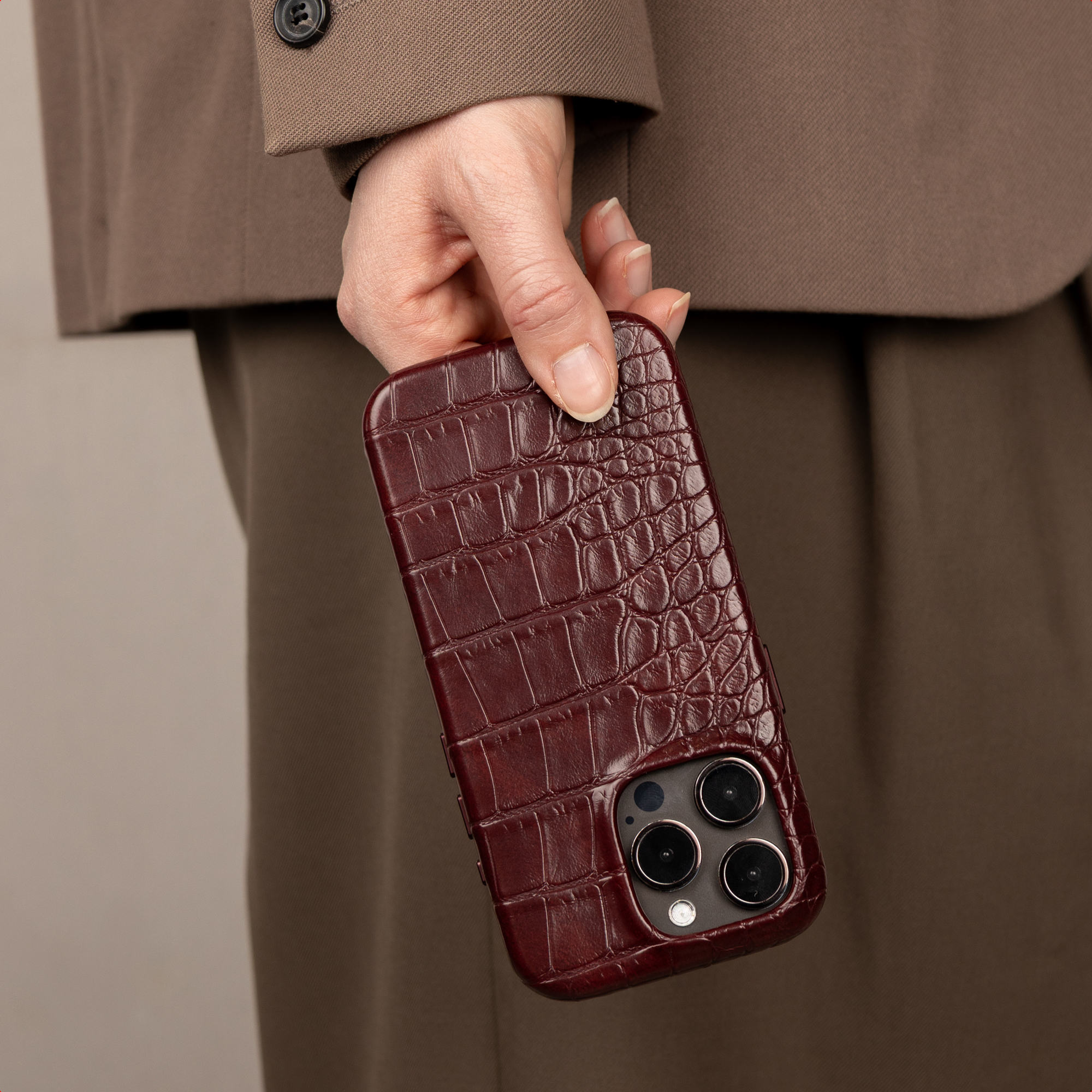 Una mano tiene uno smartphone in pelle di coccodrillo rosso scuro con abiti marroni.