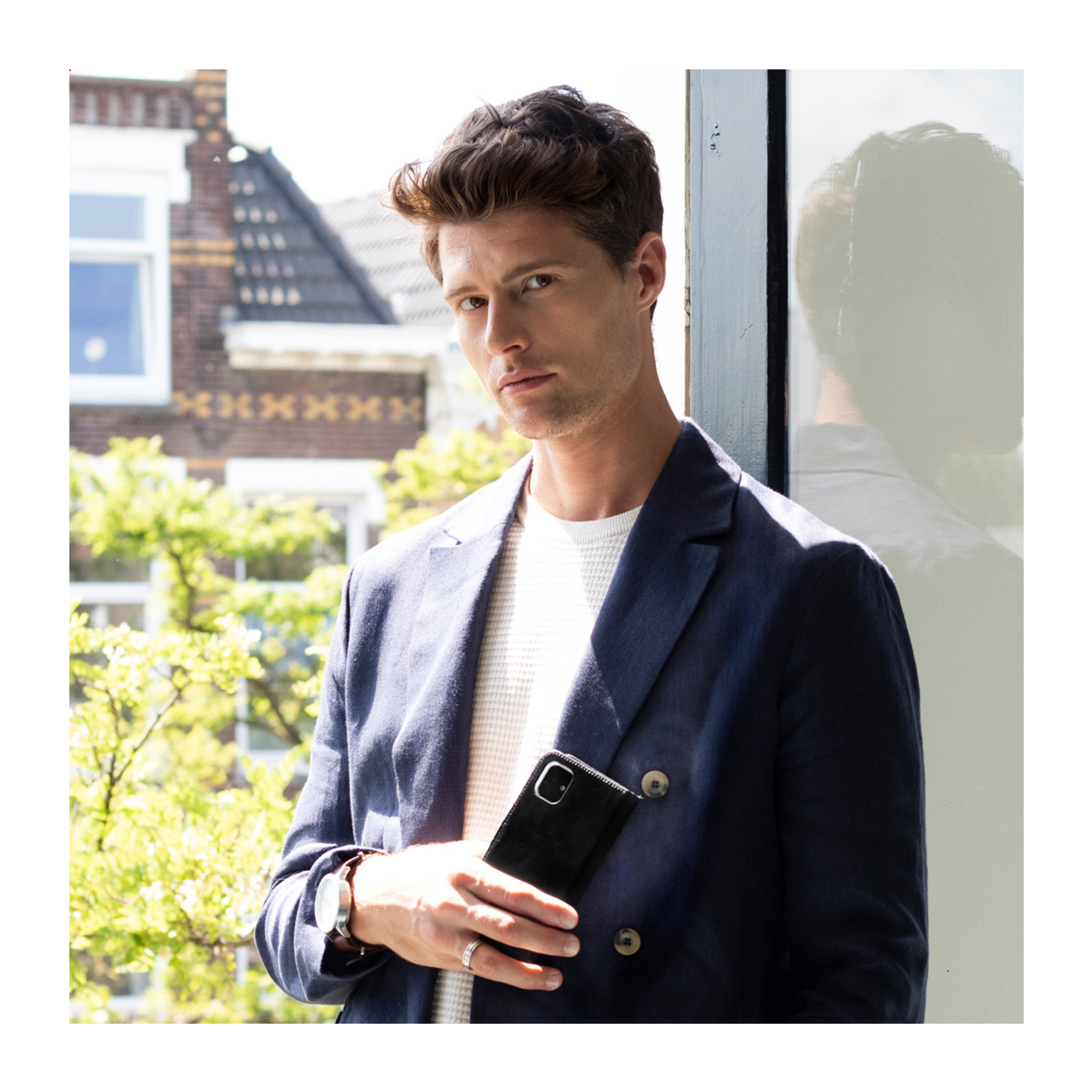 Ein Mann im blauen Sakko hält ein Smartphone, mit Gebäuden und Bäumen im Hintergrund.