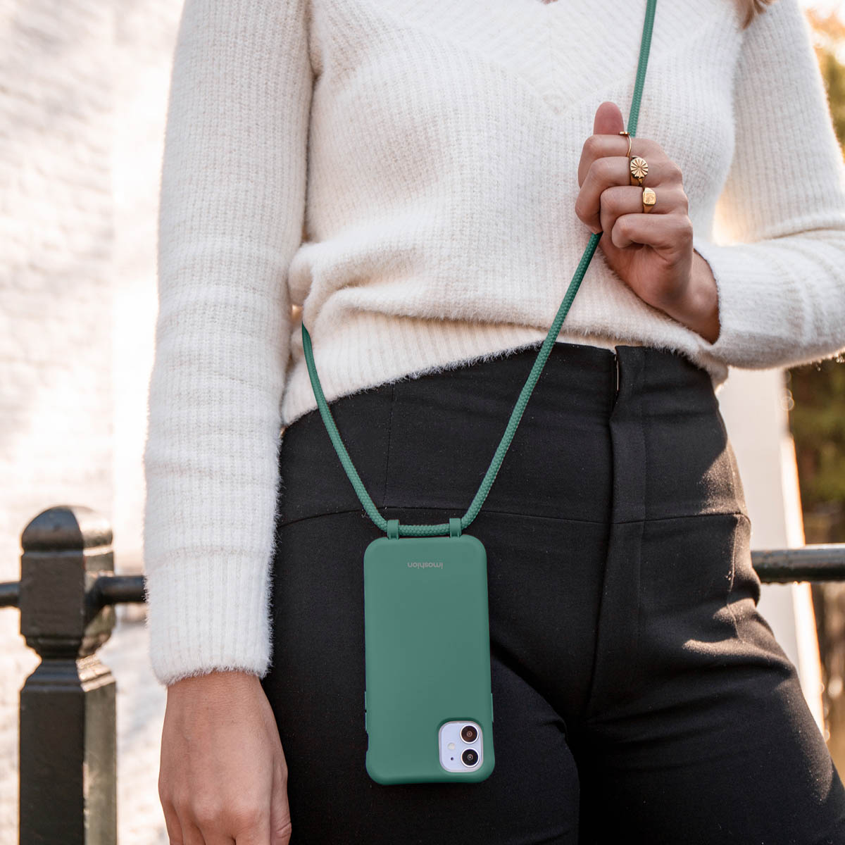 Une femme en pull blanc et pantalon noir porte un smartphone vert en bandoulière.