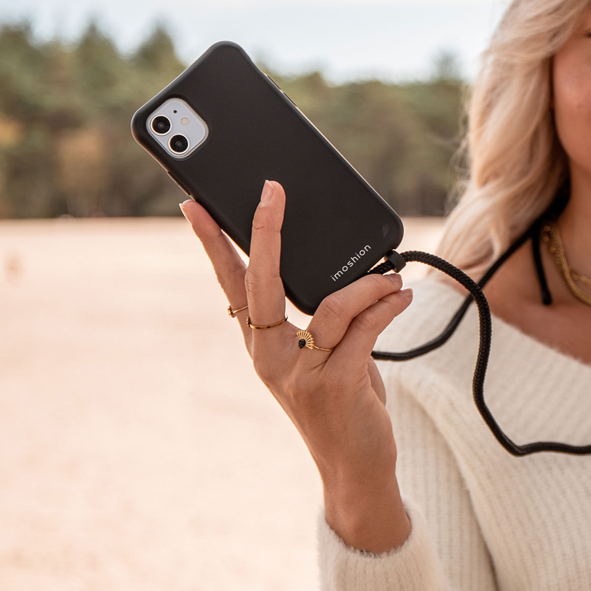Une femme en pull blanc tient un smartphone noir avec une sangle noire sur une plage de sable.