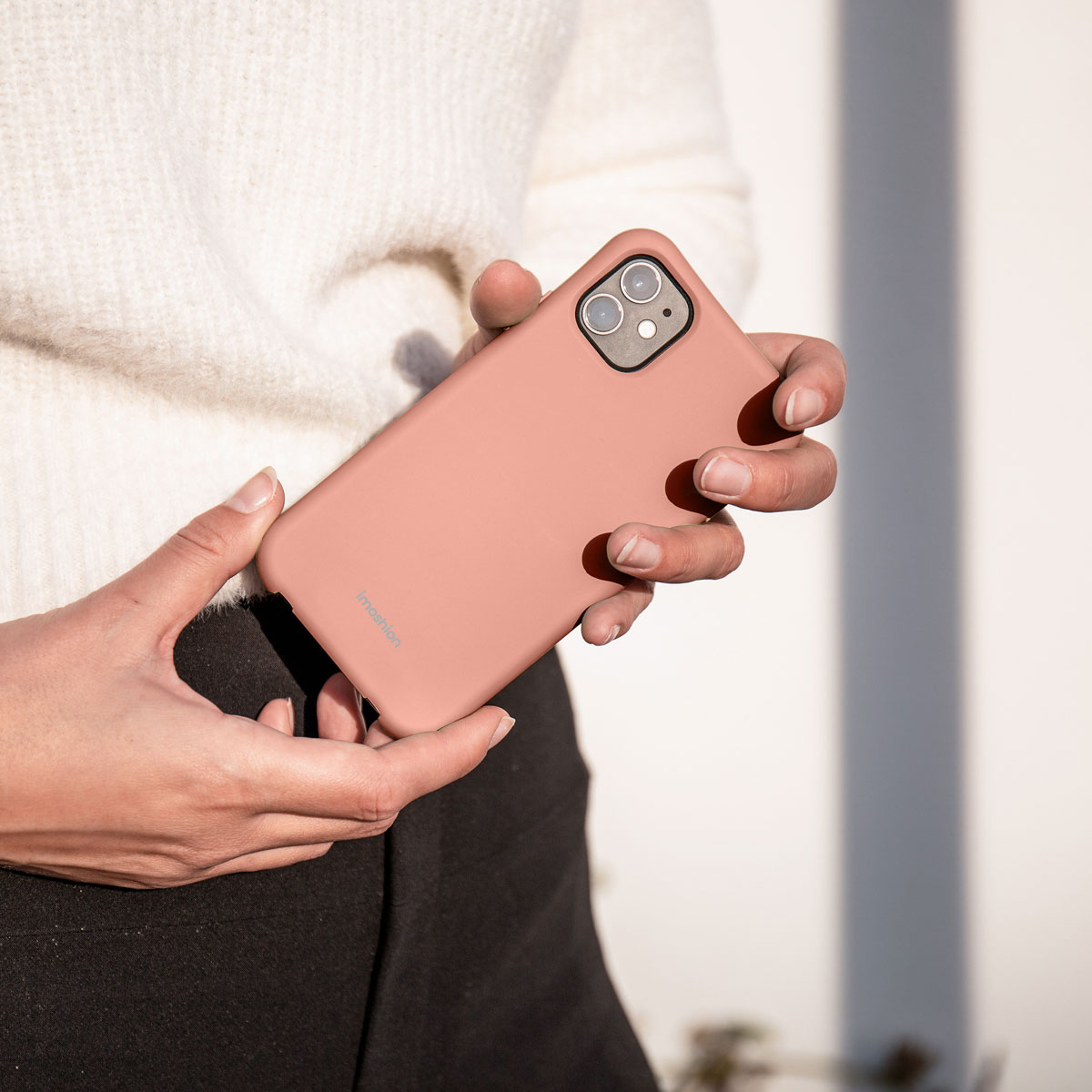 Une personne en pull blanc et pantalon noir tient un smartphone rose dans une coque en silicone.