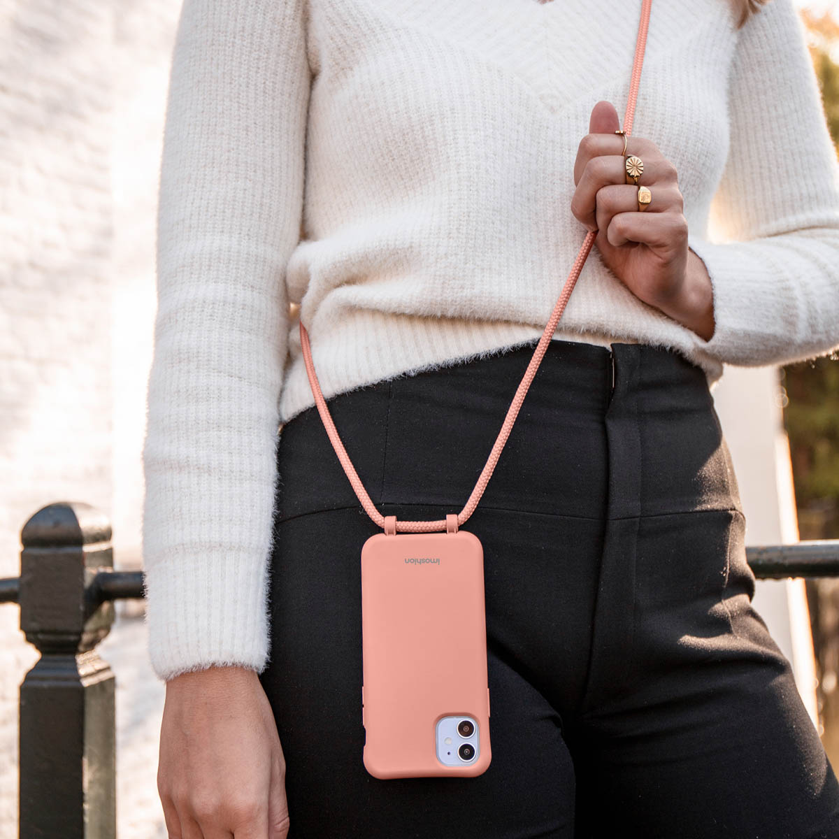 Une personne porte un pull blanc, un pantalon noir et une coque de téléphone pêche avec lanière.
