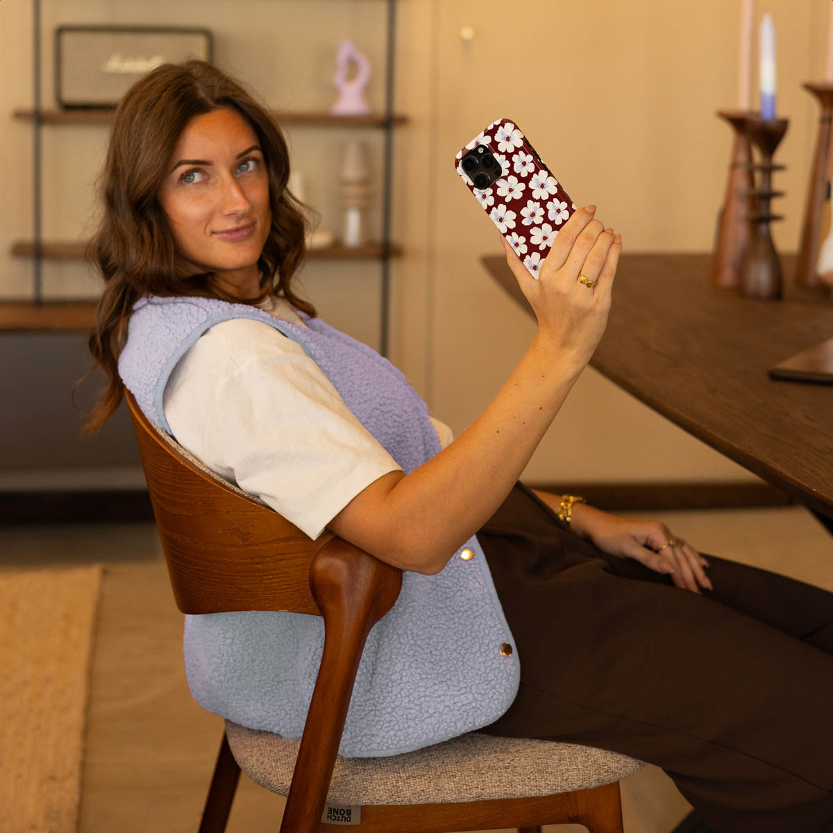 Una donna siede a un tavolo di legno reggendo uno smartphone con cover floreale, indossando maglietta bianca e gilet azzurro.