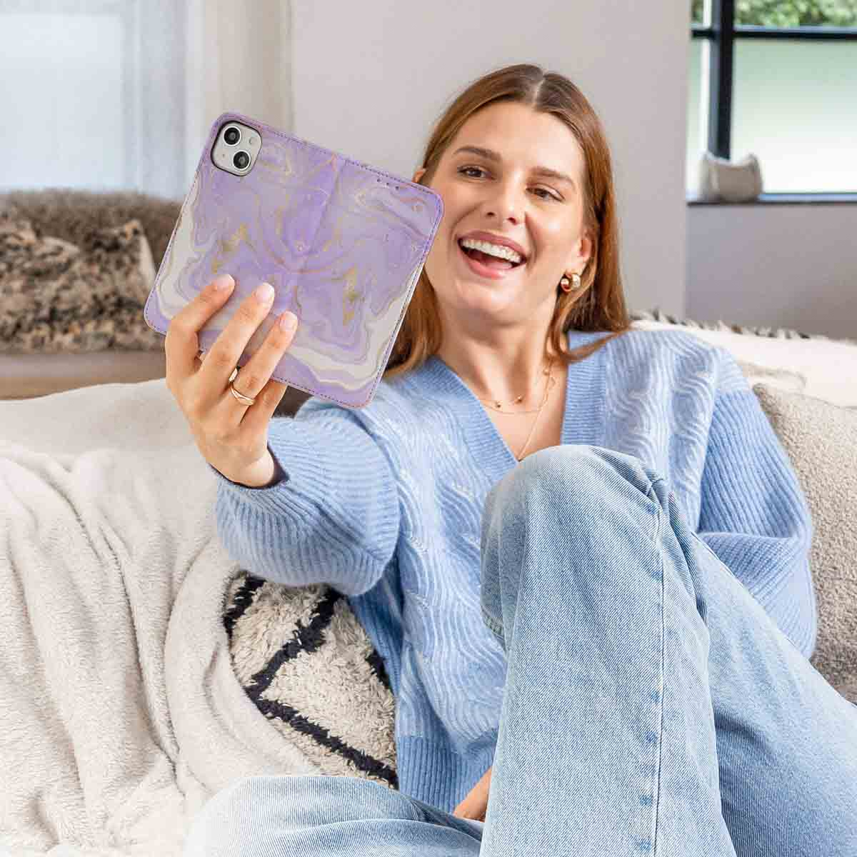 Una mujer sonriente con suéter azul y jeans sostiene una funda de teléfono morada con mármol para una selfie.