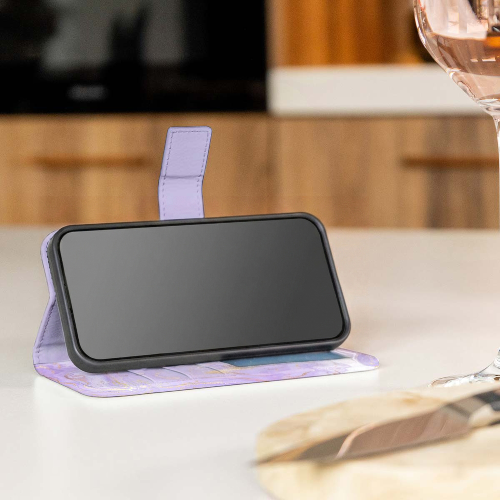 Un smartphone en una funda tipo billetera de mármol morado sobre una encimera de cocina con una copa de vino.
