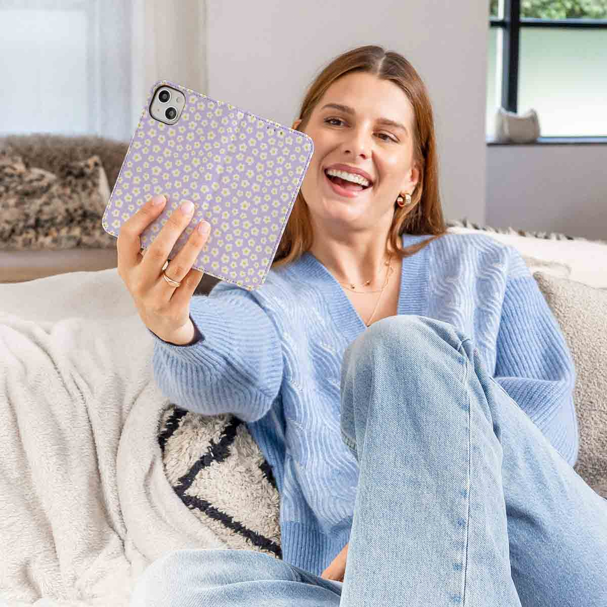 Una mujer sonriente está sentada en un sofá sosteniendo una tableta morada con patrón floral para una selfi.