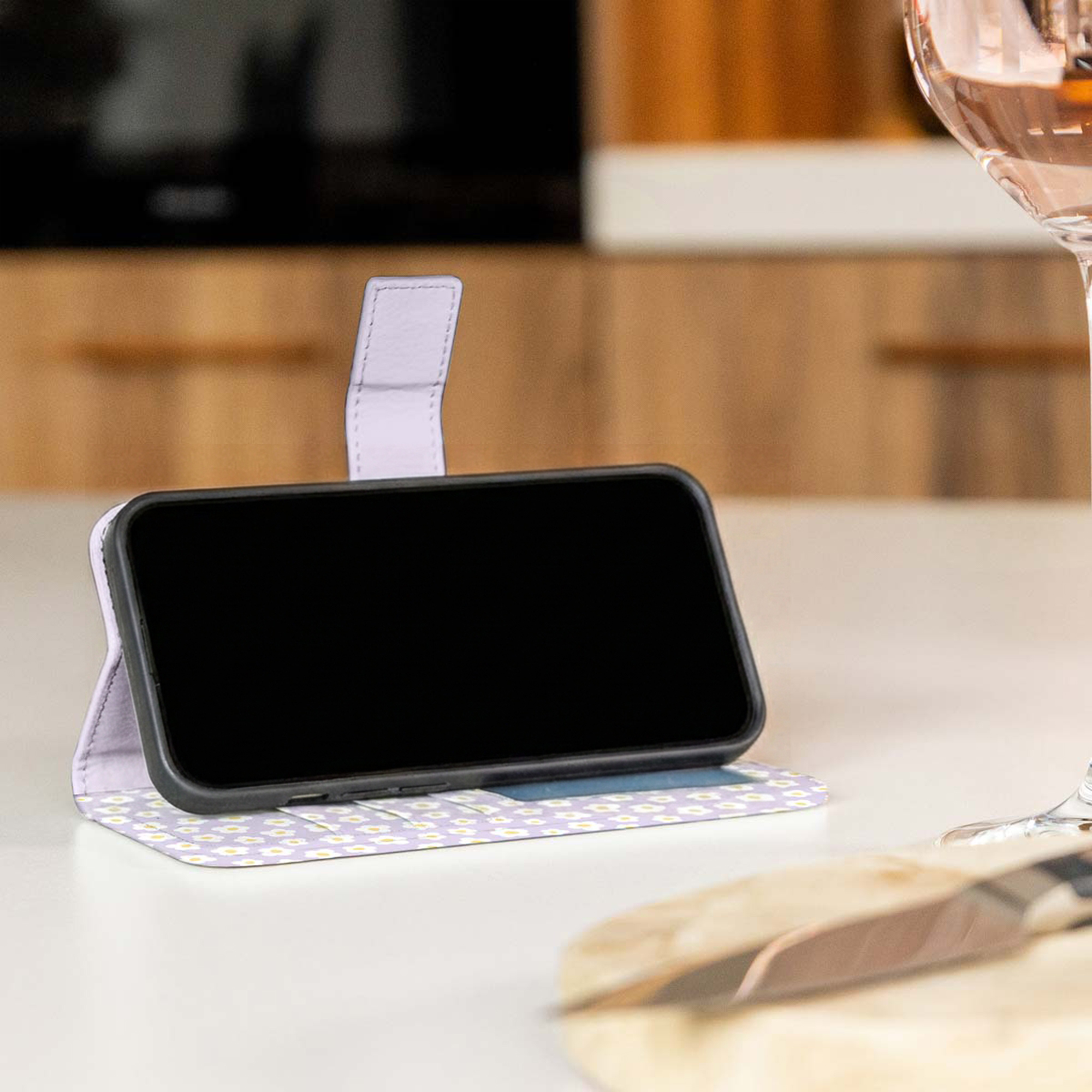 Un smartphone en una funda tipo cartera floral morada se encuentra en una encimera blanca, con una copa y cuchillo.