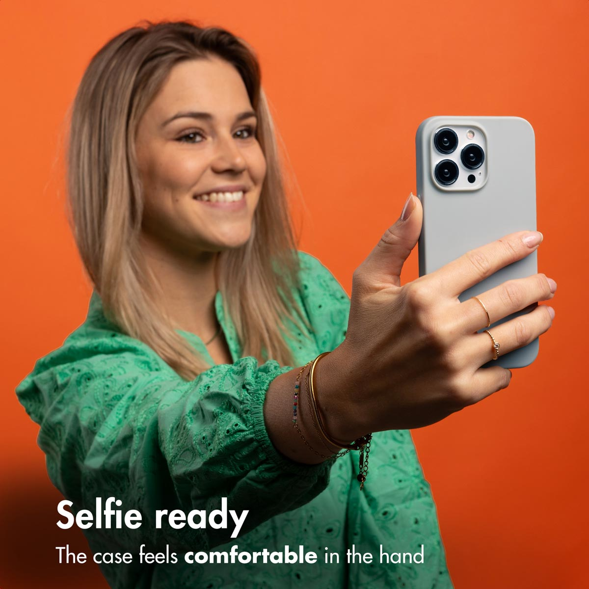 Una mujer rubia sonriente con top verde sostiene un móvil gris para un selfi ante un fondo naranja.