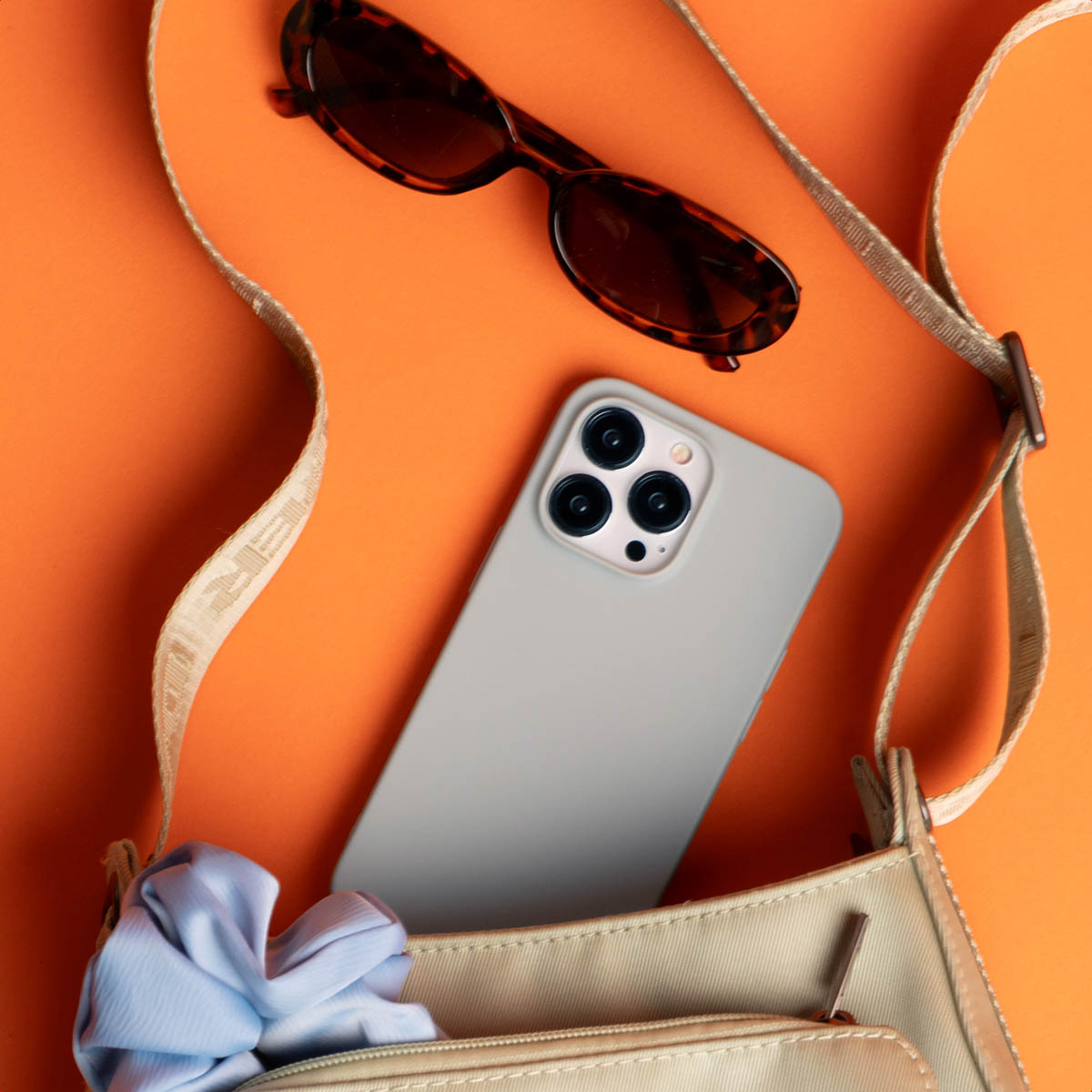 Un bolso beige, teléfono, gafas de sol y tela azul claro sobre fondo naranja.