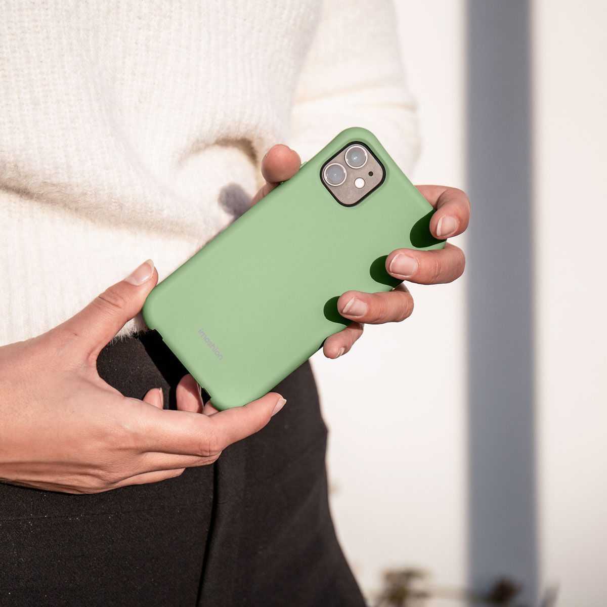 Une personne portant un pull blanc tient un smartphone avec une coque de protection verte.