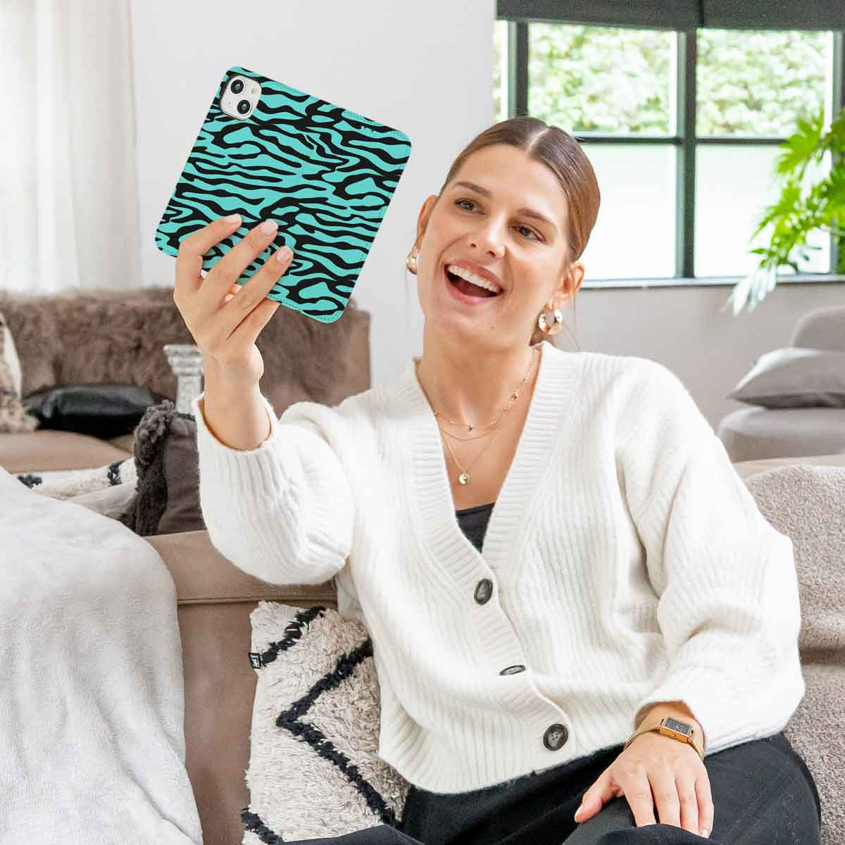 Une femme souriante en cardigan blanc prend un selfie avec une coque de téléphone zébrée turquoise.