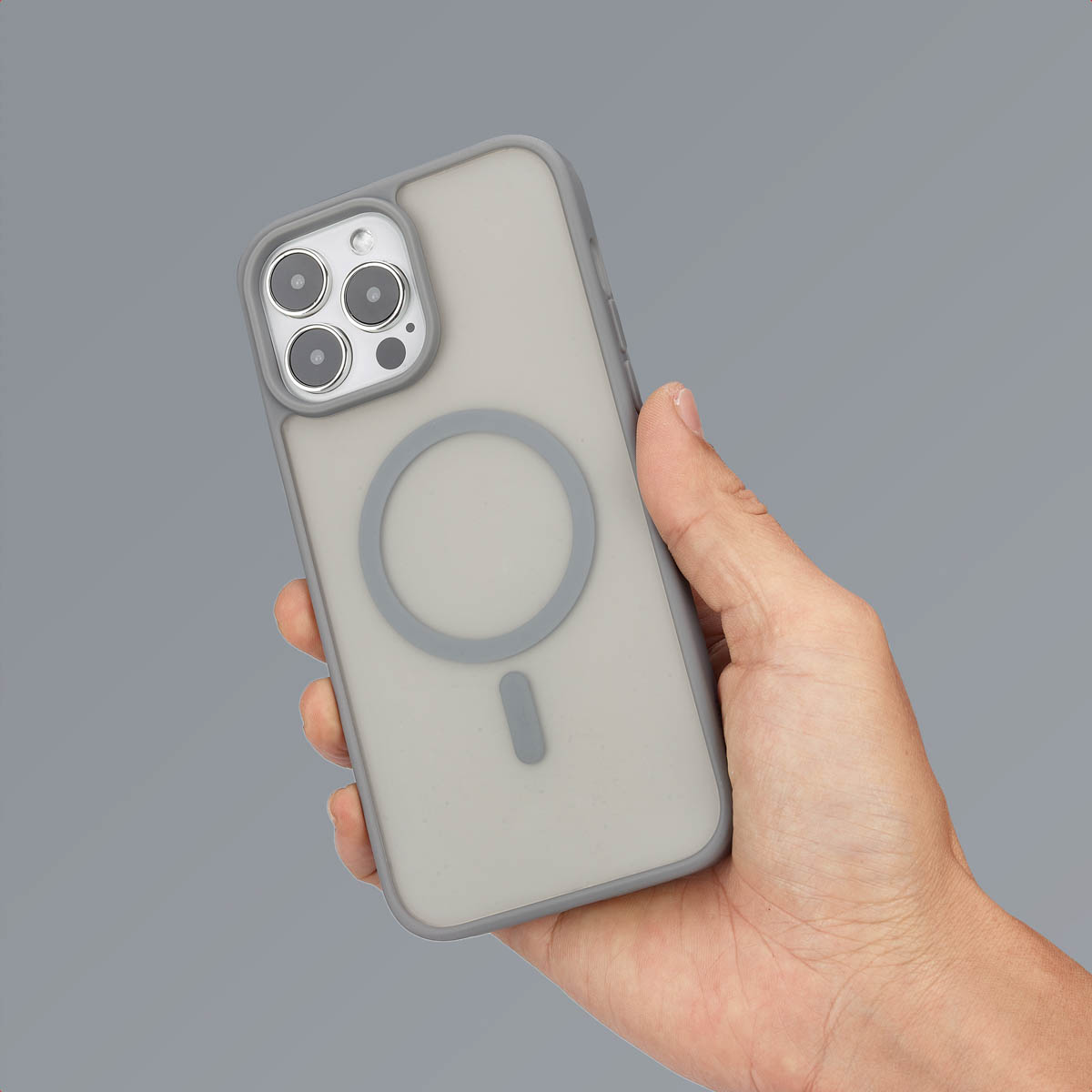 Une main tient un smartphone gris avec coque MagSafe, montrant le dos à triple caméra sur fond gris uni.