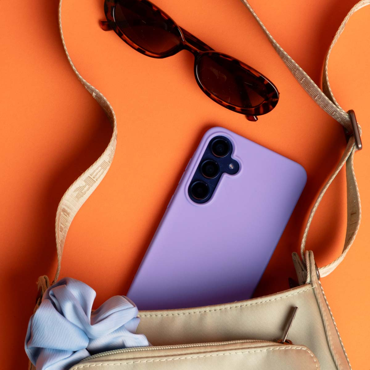 Smartphone violet dans un sac beige avec des lunettes de soleil et un chouchou bleu sur fond orange.
