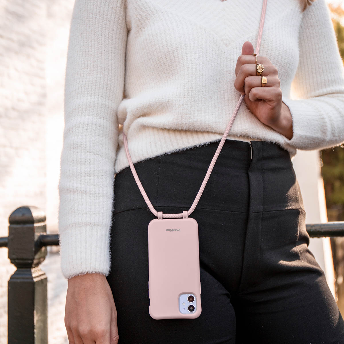 Une femme en pull blanc et pantalon noir porte un smartphone rose avec une lanière.