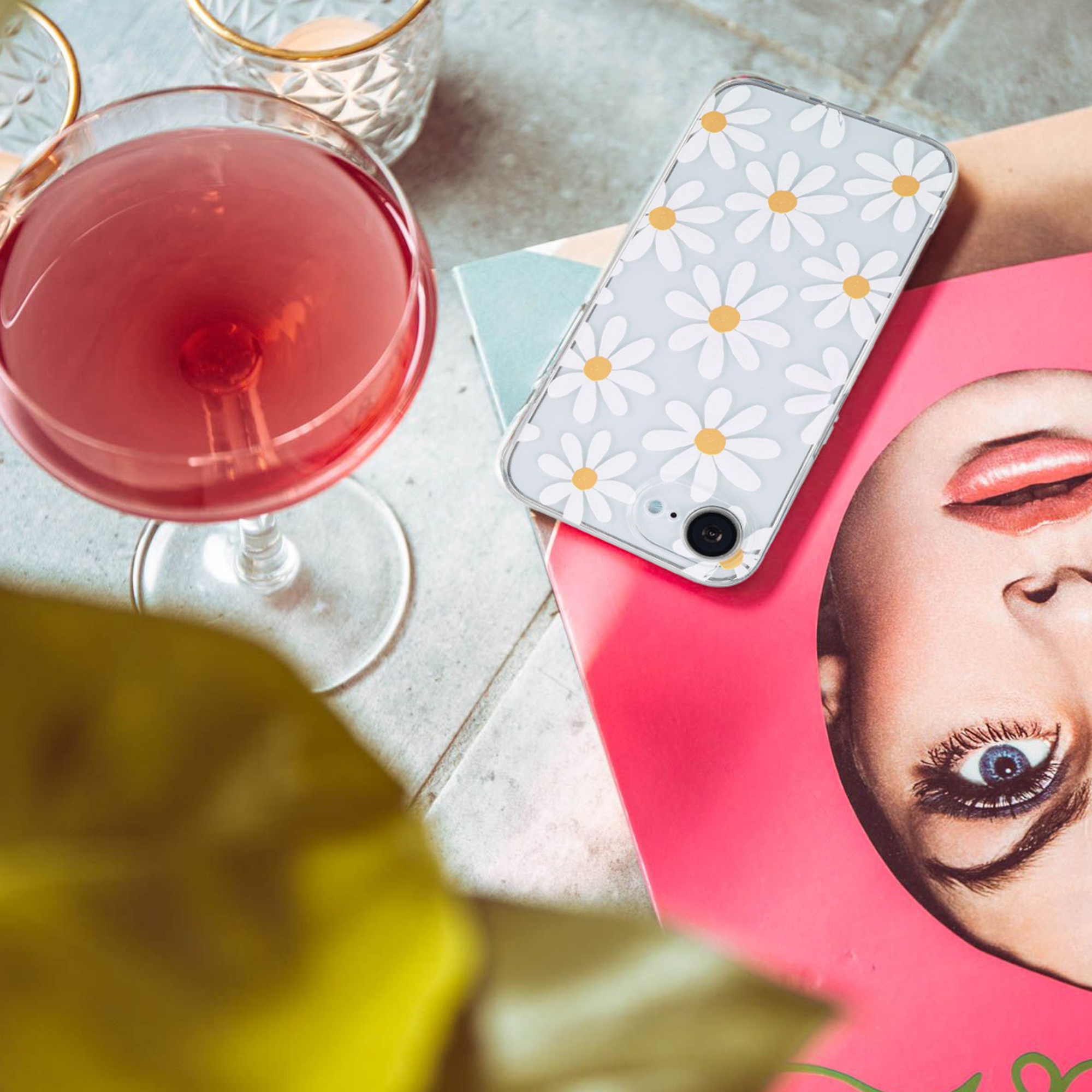 Un smartphone avec coque marguerites, un verre de cocktail rouge et un magazine sur une table.