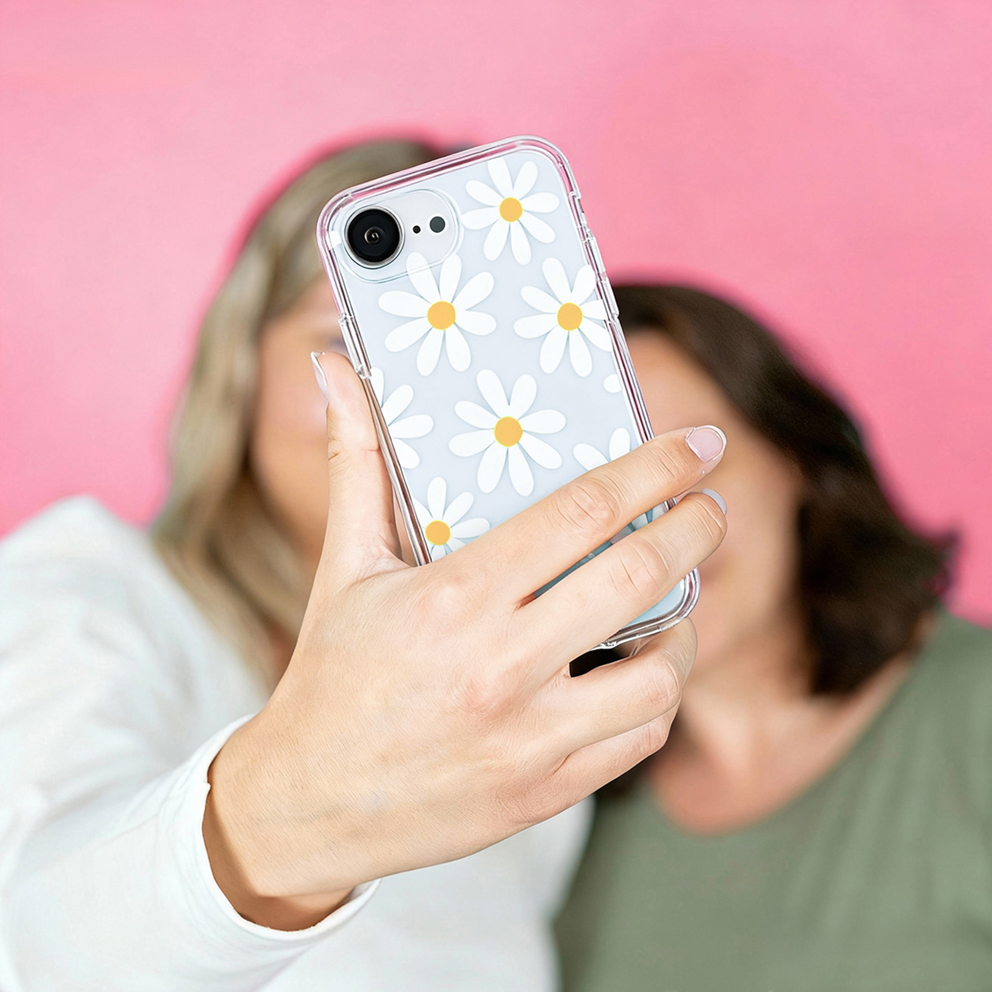Une main tient un téléphone à motif marguerites, deux femmes floues en arrière-plan devant un mur rose.