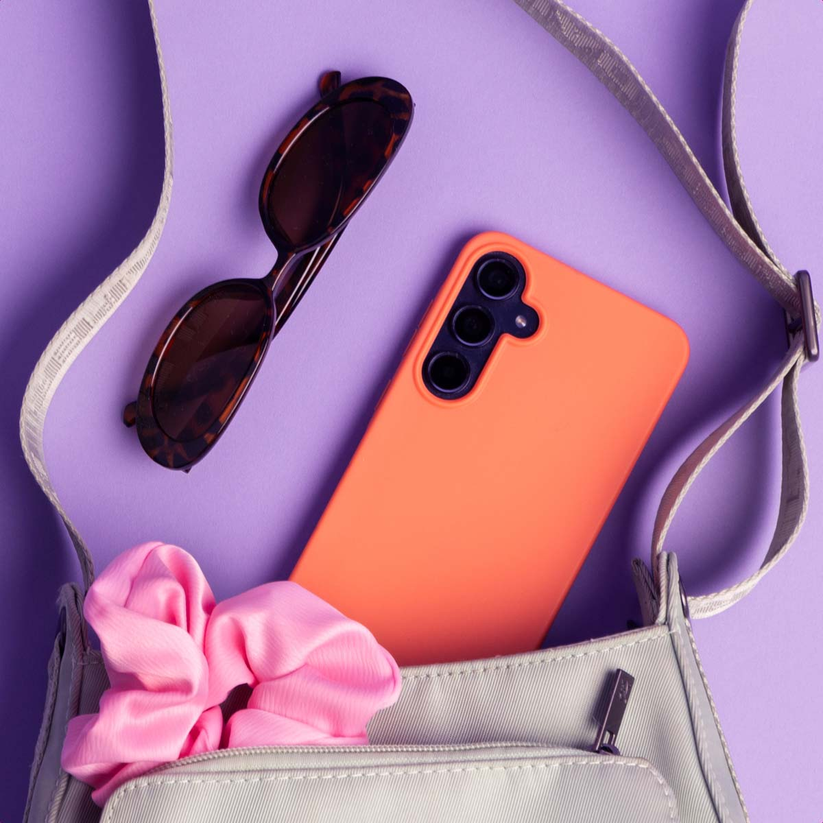 Un teléfono naranja, un coletero rosa y gafas de sol carey están en un fondo morado junto a un bolso.