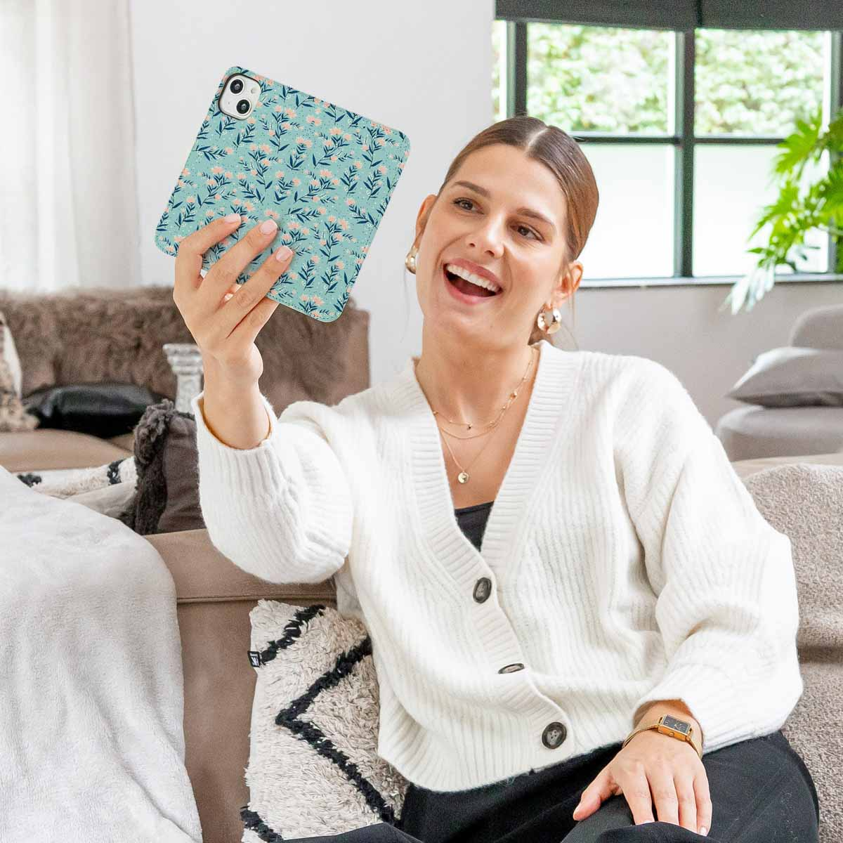 Una mujer sonriente se sienta en un sofá, levantando una tableta con funda de flores azules para tomarse un selfi.