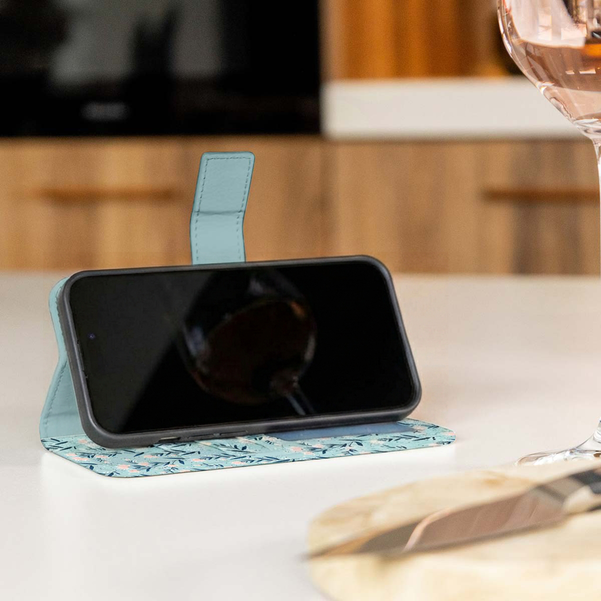 Un teléfono en una funda con patrón floral azul claro sobre una mesa blanca junto a una copa de vino.