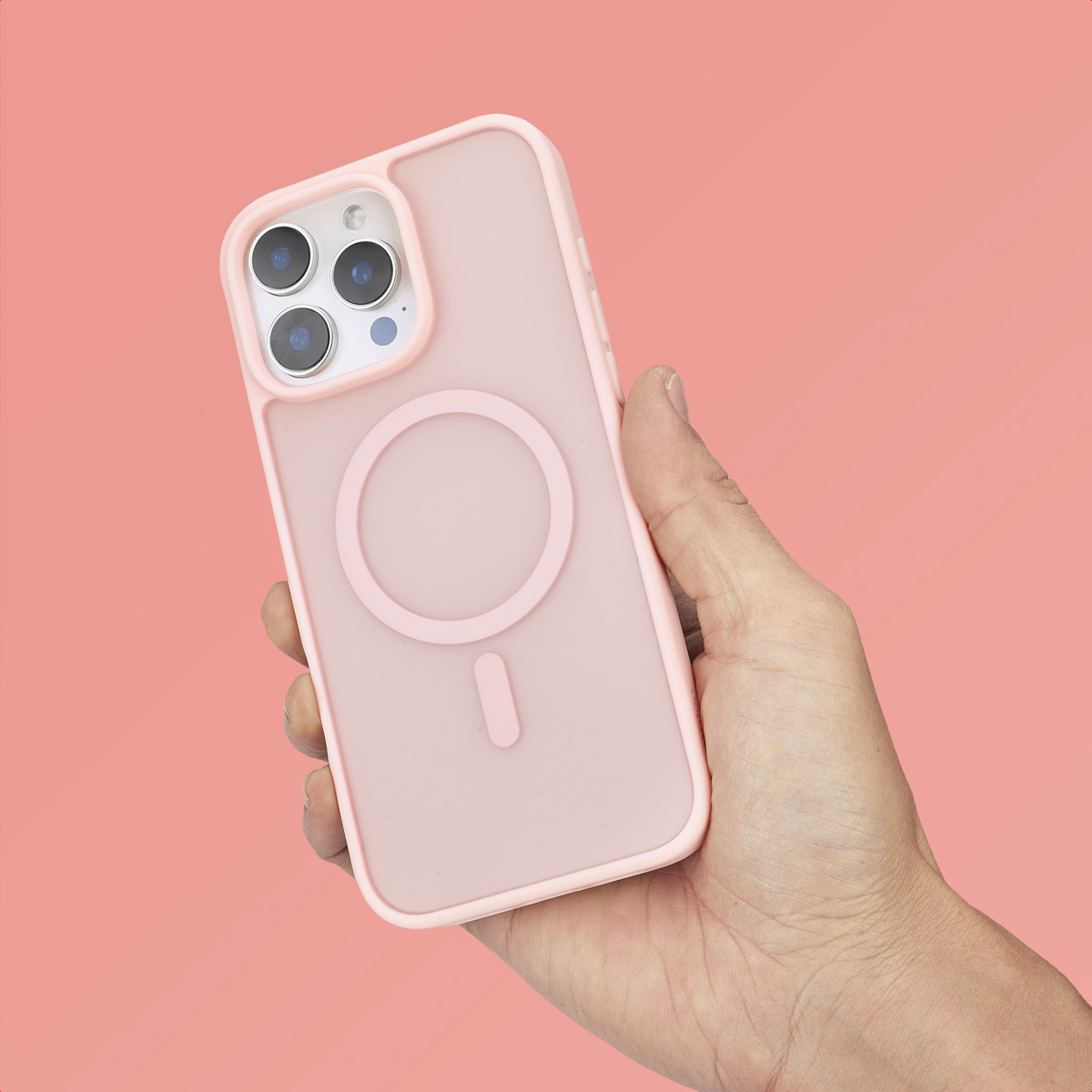 Una mano sostiene un smartphone rosa con funda protectora y anillo MagSafe sobre fondo rosa.