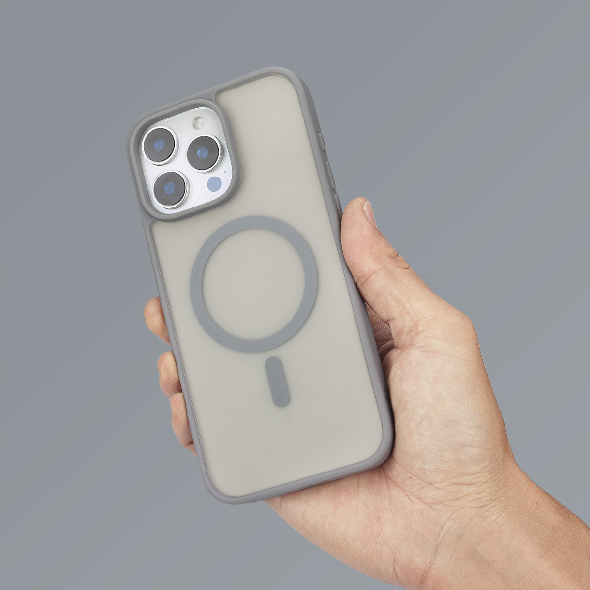 Una mano sostiene un teléfono gris con funda transparente y anillo MagSafe.