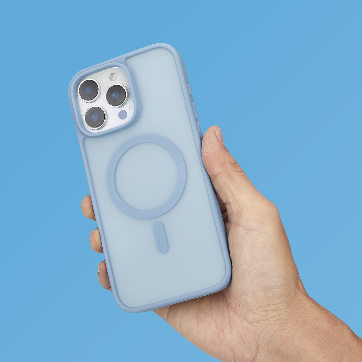 Una mano sostiene un smartphone azul claro con funda a juego y anillo Magsafe sobre fondo azul.