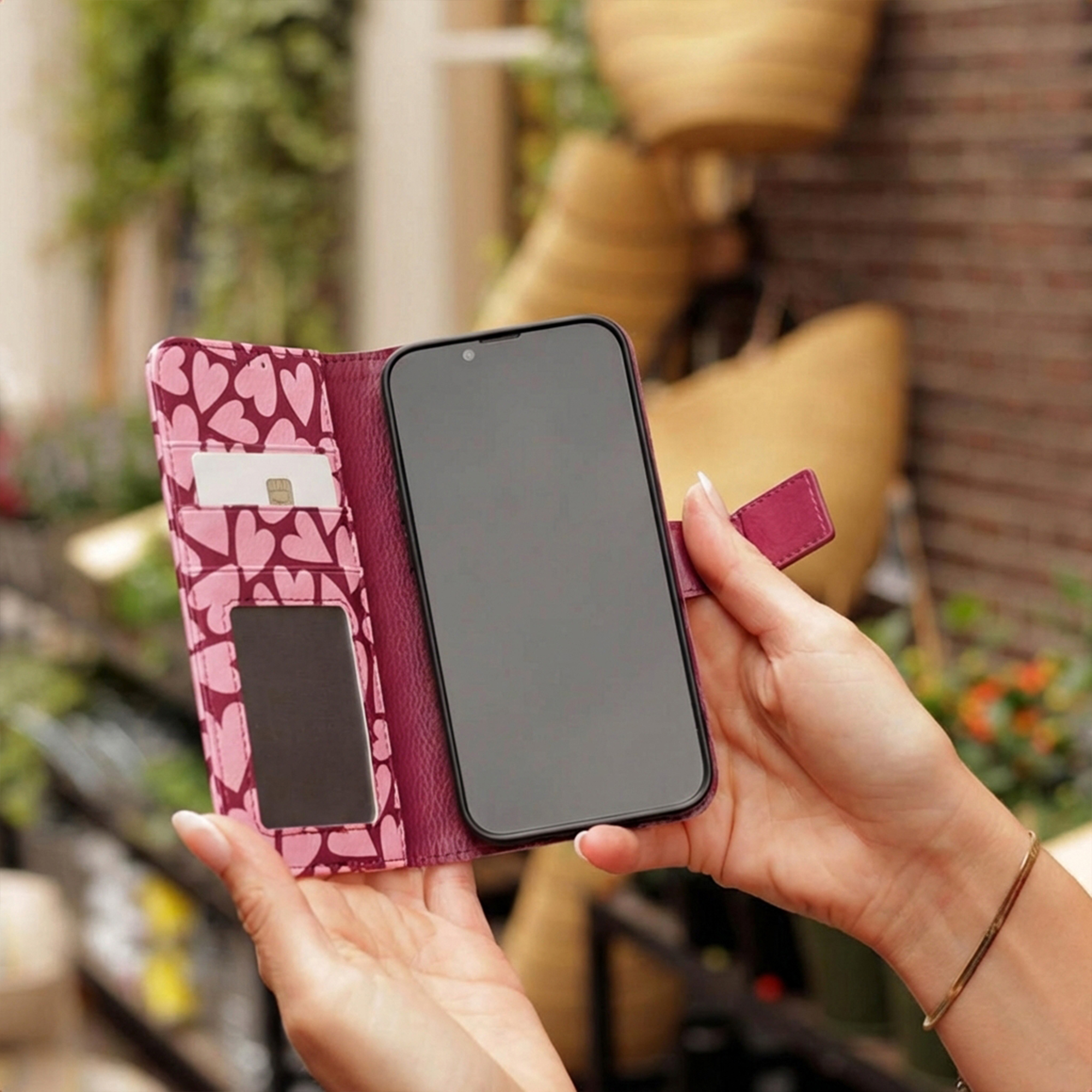Eine Hand hält eine pinkfarbene Handyhülle mit Herzmustern, Kartenfächern und einem Smartphone.