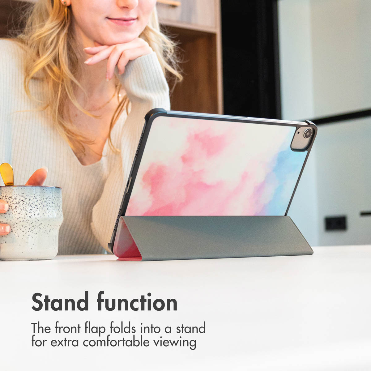 A blonde woman sits at a table with a tablet in a grey case featuring a pink and blue design, resting on the table.