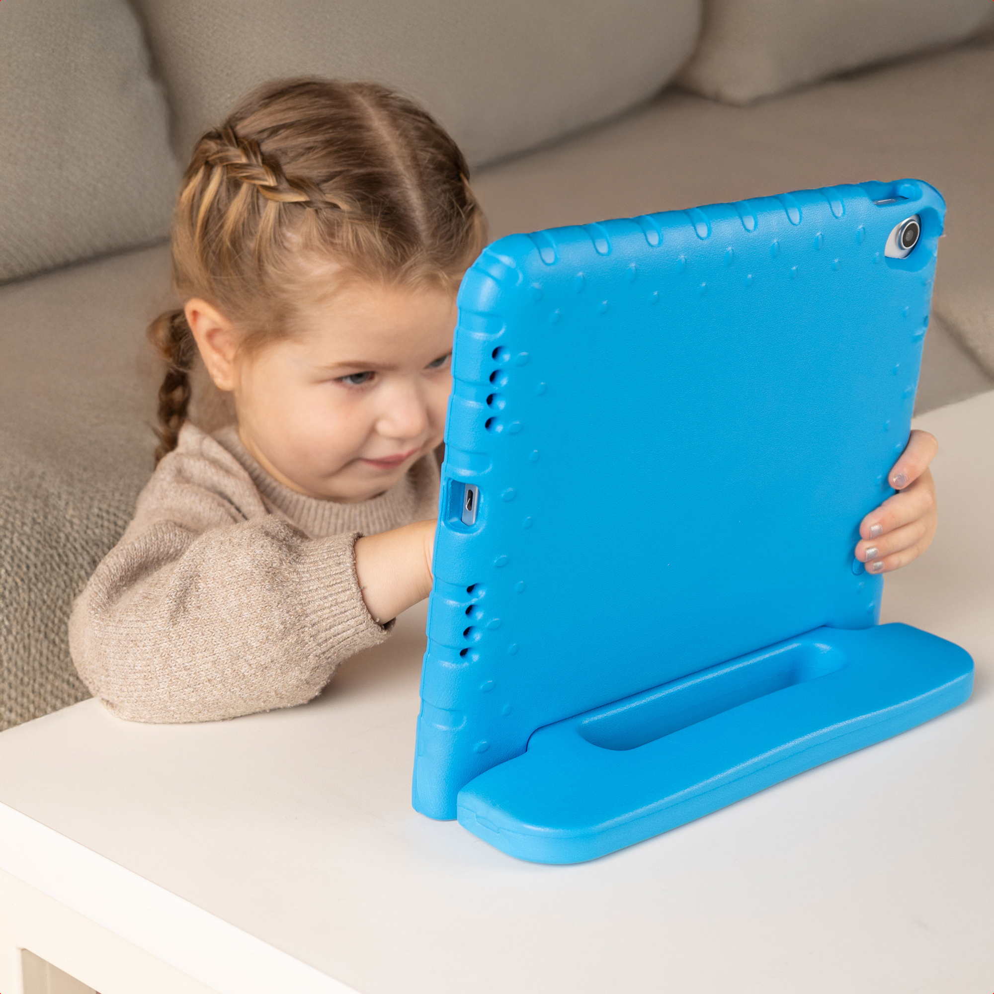 Una bambina con un maglione beige siede a un tavolo reggendo un tablet con una custodia protettiva blu.