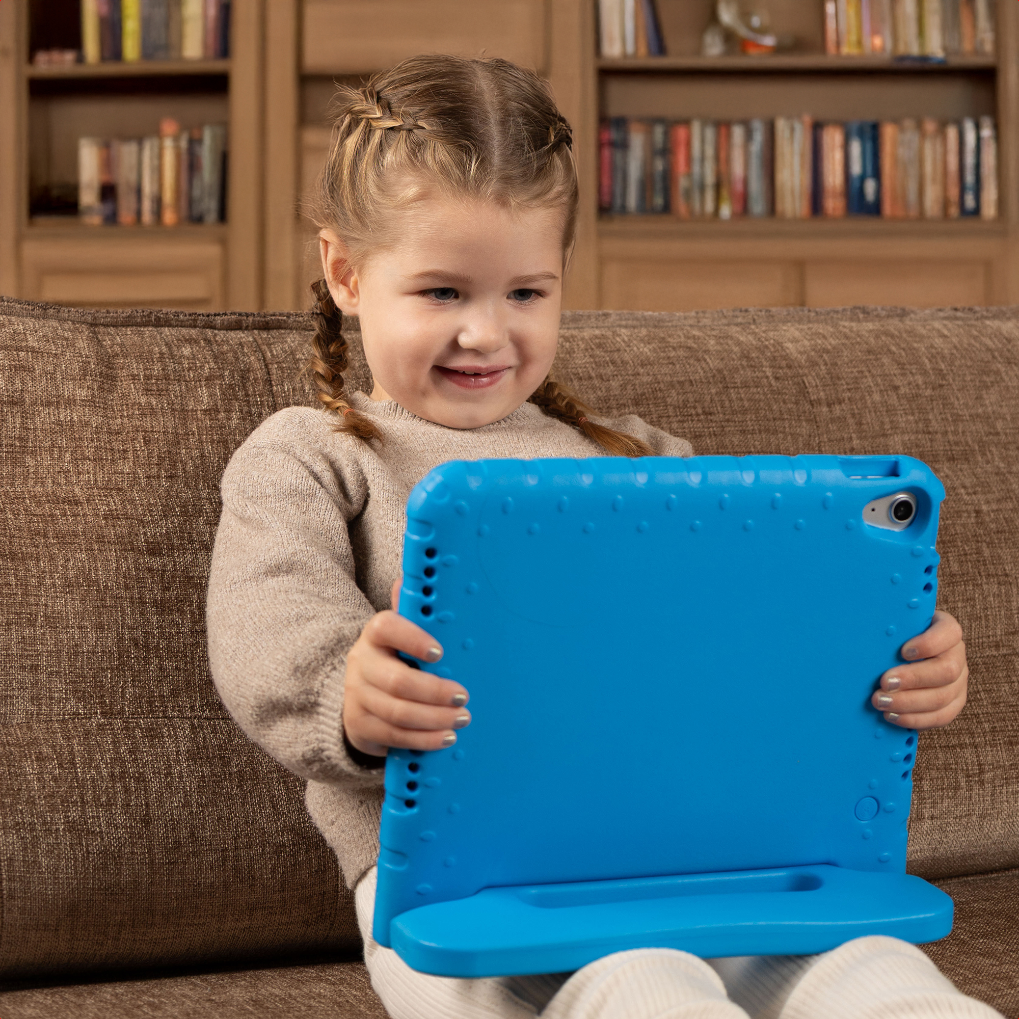 Una bambina con i capelli intrecciati sorride tenendo un tablet blu su un divano con librerie sullo sfondo.