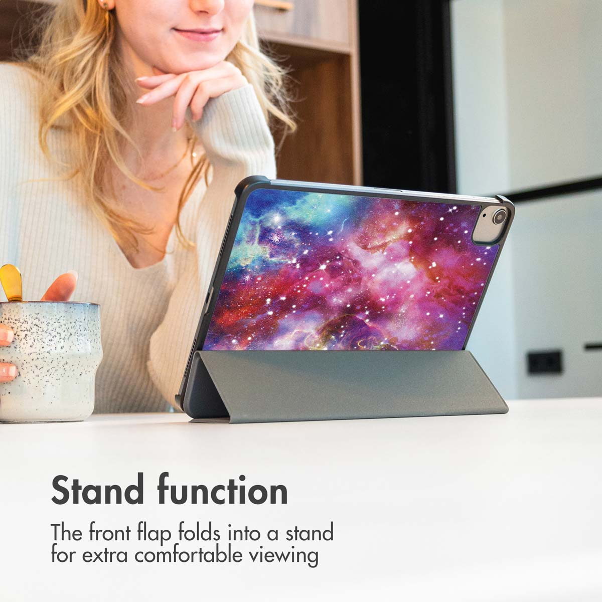 Une femme blonde est assise à une table avec une tablette dans un étui design galaxie.